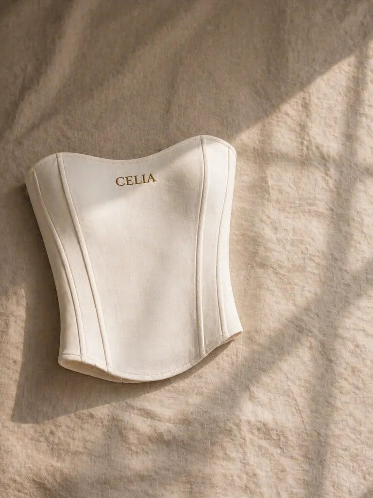 Editorial still-life photograph, natural light. A single white CELIA corset garment rests on a textu
